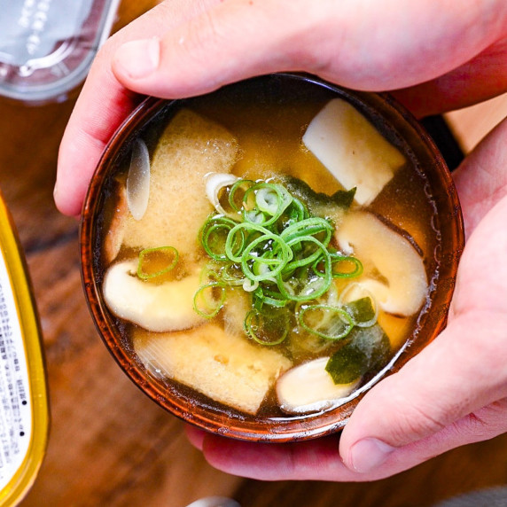A wooden bowl of miso soup made with shiitake mushrooms, tofu and wakame seaweed held by two hands
