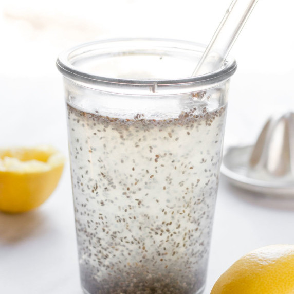 A clear glass of chia seed drink with a glass straw sits with fresh lemons around it.