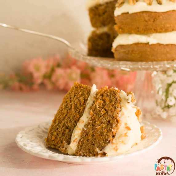 A slice of naked carrot cake on a white plate with flowers and cake in the background.