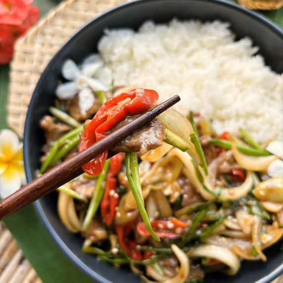Nua pad prik, a Thai chili beef stir-fry, lifted by a pair of chopsticks over a black bowl.