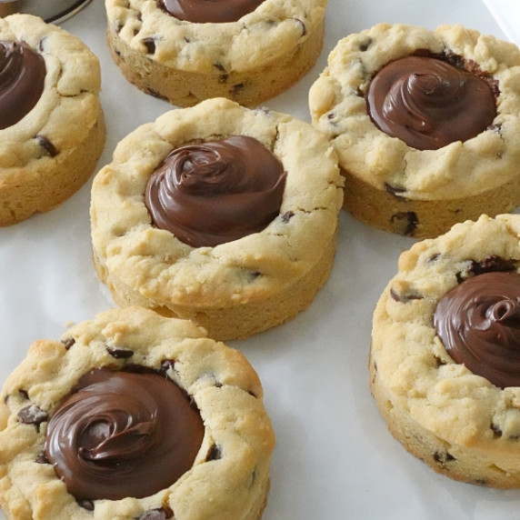 Thick chocolate chip cookie cups filled with creamy Nutella on a white tray.