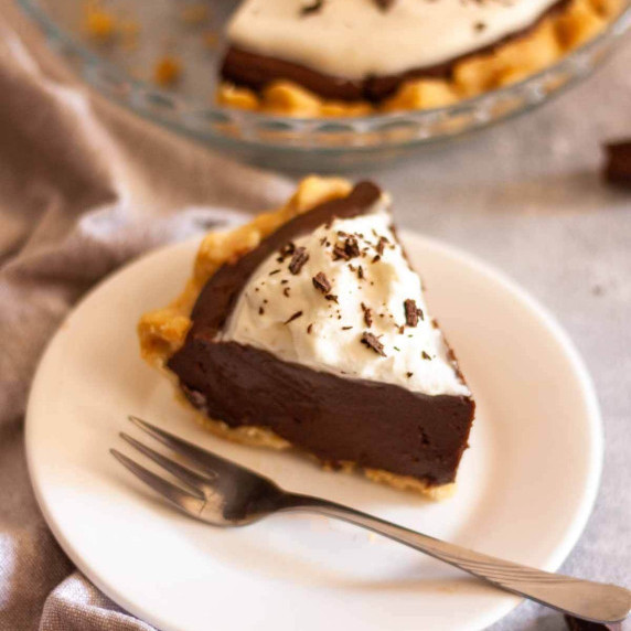 A slice of chocolate pie on a white plate in front of a pie dish with chocolate pie.