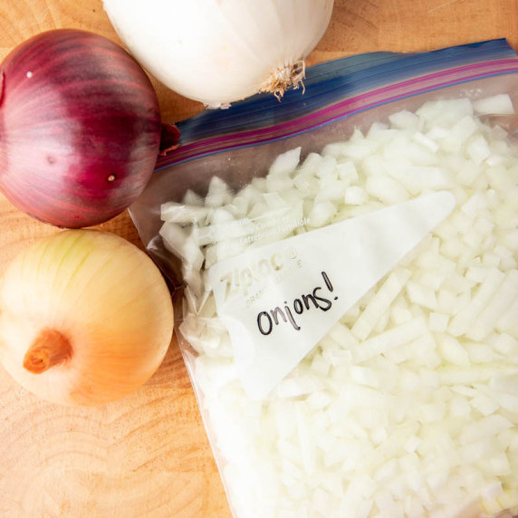 A freezer bag filled with chopped onions sits with a red, yellow, and white onion beside it.