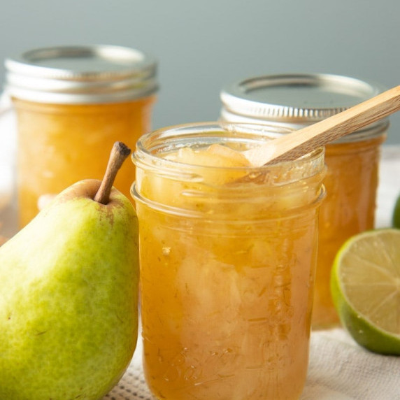 An open jar of ginger pear preserves stands with a pear leaning on it and a spoon scooping into it.
