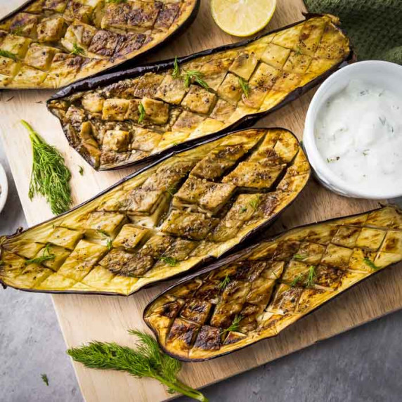 Overhead shot of four slices of roasted eggplant on a wooden cutting board with fresh dill.