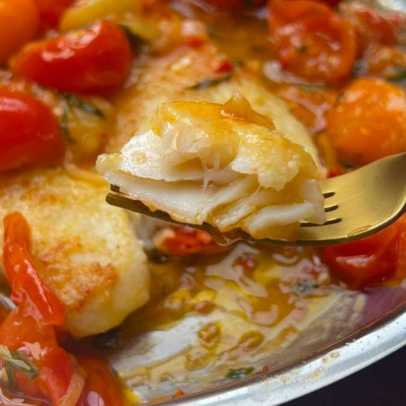 a piece of cooked fish on a fork with fish in cherry tomato sauce in the background on a pan