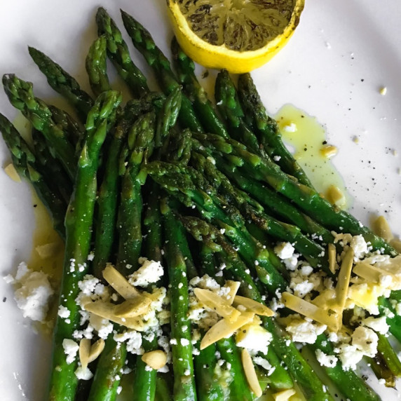 pan charred asparagus on a white plate with feta, almonds, lemon dressing and charred lemon half.