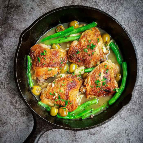 Overhead shot of pan fried chicken thighs with olives and green beans in a skillet on a black table.