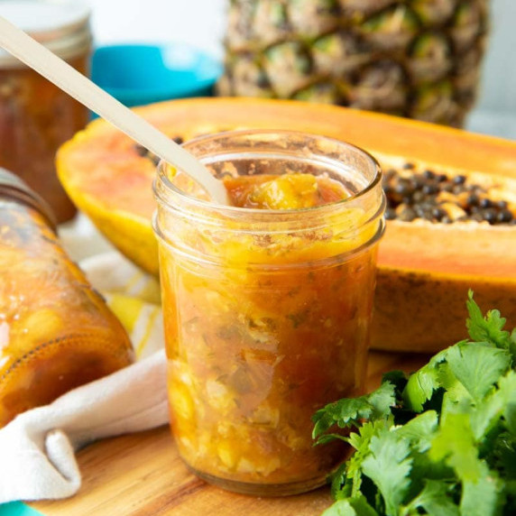 A spoon dips into a jar of canned pineapple papaya chili salsa with fresh fruit and herbs around.