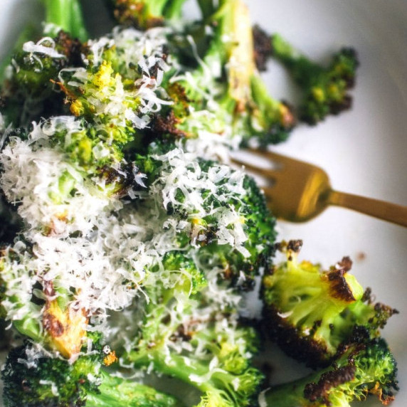 A gold fork rests at the bottom of a pile of parmesan roasted broccoli on a white serving dish.