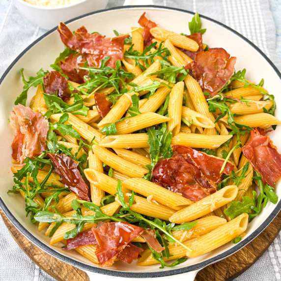 A bowl of penne pasta mixed with arugula and crispy prosciutto. 