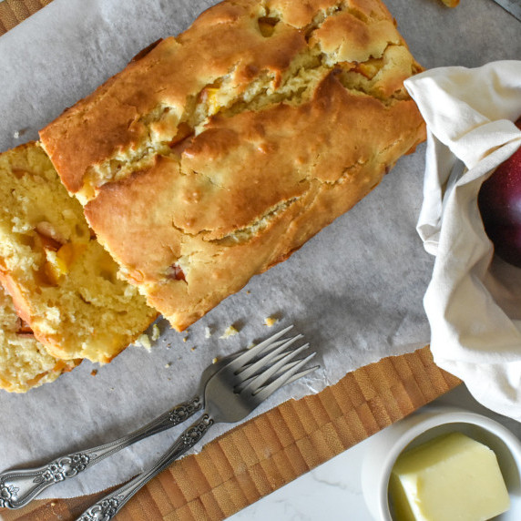 Sliced peach yogurt loaf cake on a cutting board with forks, butter, fruit on the side.