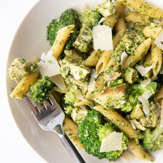 A fork rests in a bowl of pasta holding a bit of pesto penne, chicken, and broccoli.