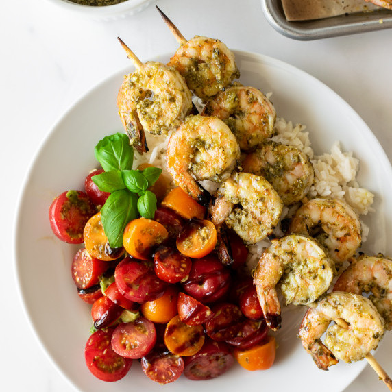 pesto shrimp on skewers over rice with tomatoes garnished with basil in a white bowl.