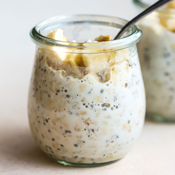 Small glass jar with protein overnight oats topped with banana slices on beige and white surface