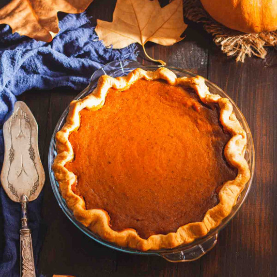 Pumpkin pie in a glass pie dish on a wooden surface next to a pumpkin and autumn leaves.