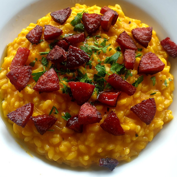 a white plate with pumpkin risotto, garnished with parsley and crunchy sausage.