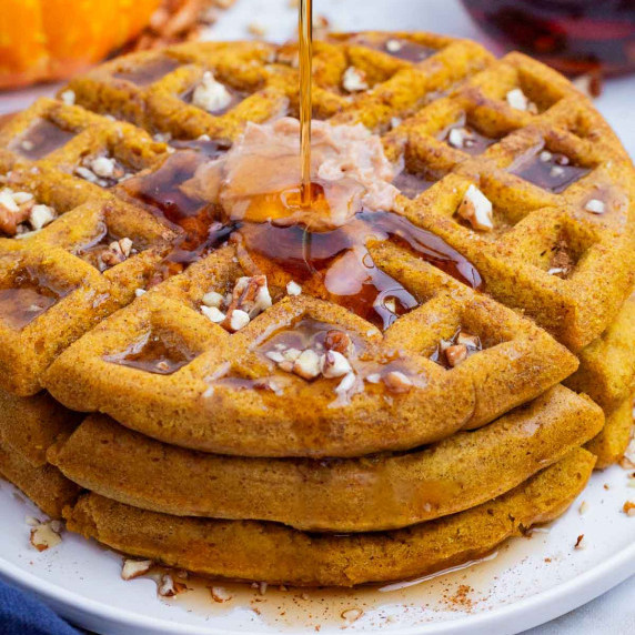 Pumpkin Waffles RECIPE stacked on a white plate with syrup being drizzled on top. 