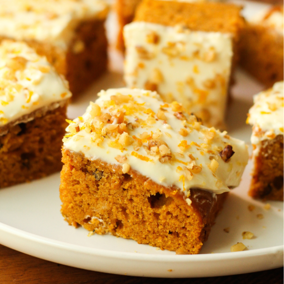 Pumpkin cake squares with orange cream cheese frosting on a white plate.