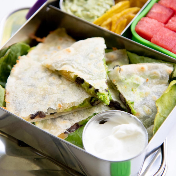 An avocado and black bean quesadilla in a metal lunch box with a small cup of sour cream for serving