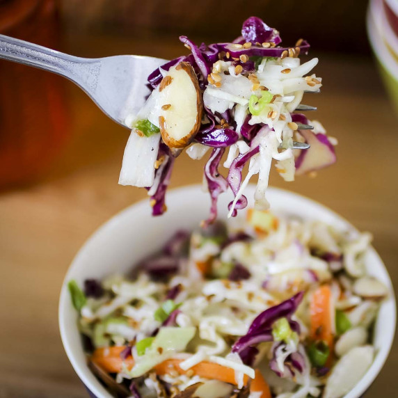 Side close up shot of a fork holding up a bite of coleslaw over a bowl of it.