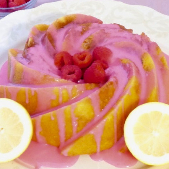A raspberry lemon cake on a white plate with sliced lemons and a bowl of raspberries.  