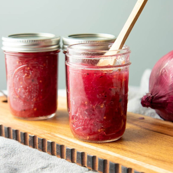 Three half-pint jars of red onion and port wine jam stand on a wooden board, a spoon scoops from one