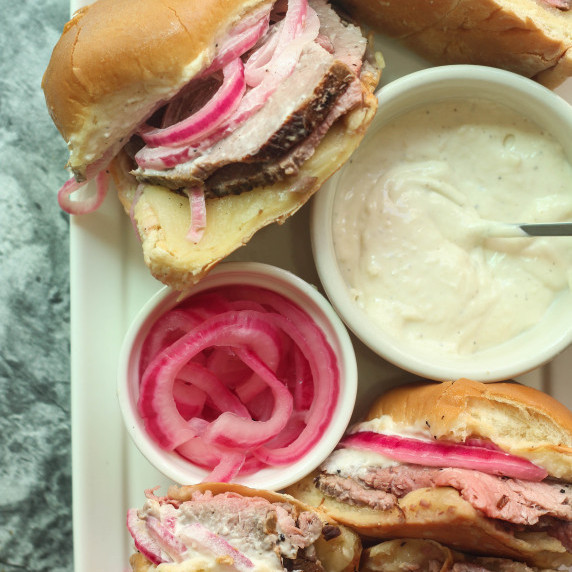 Roast beef sliders on a white plate with a bowl of garlic horseradish aioli and pickled onions.