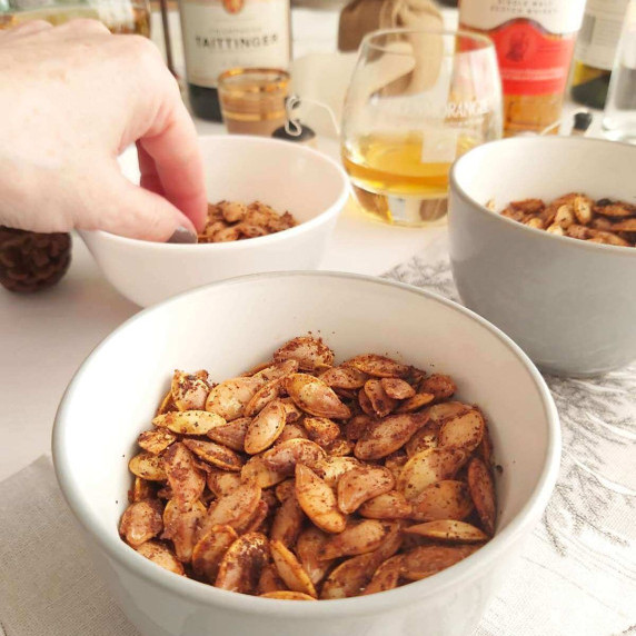 Image of party table, with bowl of seasoned pumpkin seeds, and female hand and drinks to the back.