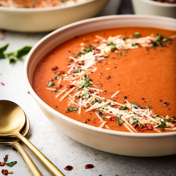 Side shot of a large white bowl with red pepper soup garnished with fresh herbs and parmesan cheese.