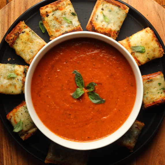 Roasted Tomato Basil and Red Pepper Soup in a white bowl, with cheesy garlic bread squares.