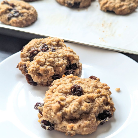 Two rum raisin oatmeal cookies on a plate with a tray of cookies in the background.