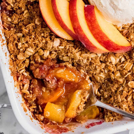A spoon dips into the corner of a fresh peach crisp in a white baking dish, topped with peach slices