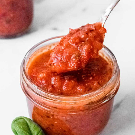 A spoon scoops roasted red pepper sauce from a small glass jar, fresh basil leaves beside it.