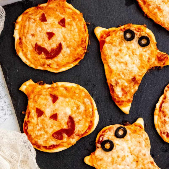 Mini pizzas shaped like jack-o-lanterns and ghosts with topping faces are served on a slate board.