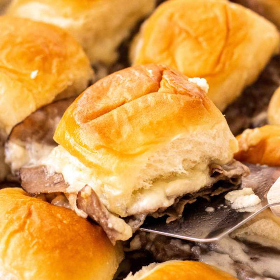 A metal spatula lifts a baked roast beef slider from the center of a baking of sliders.
