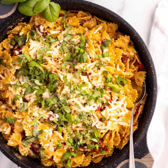 A spoon tucks into a skillet of creamy bowtie pasta with sun-dried tomatoes garnished with basil.