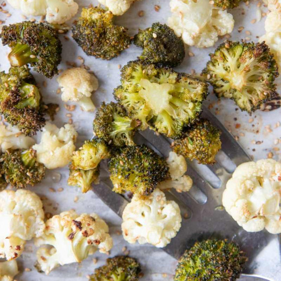 A metal spatula slides under roasted broccoli and cauliflower on a parchment lined sheet pan.