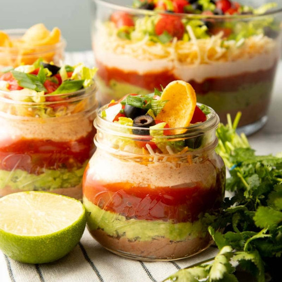 Two individual servings of seven layer dip in glass jars sit in front of a large serving bowl of dip