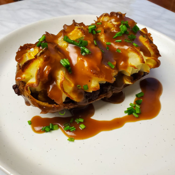 Crispy potato skin stuffed and topped with piped mash & gravy on a white plate.