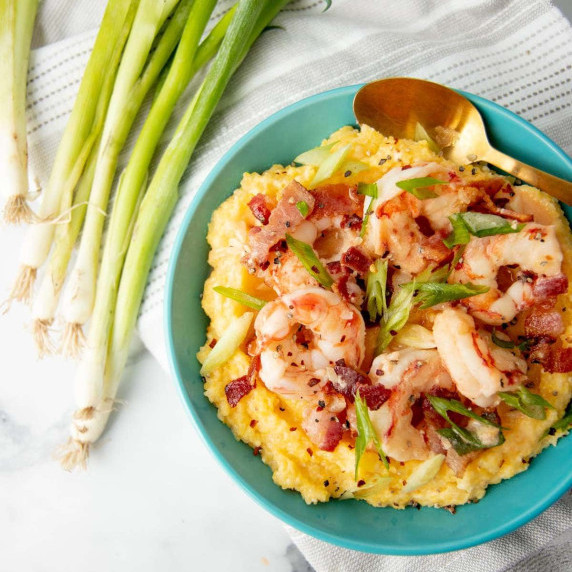 A single-serving of shrimp and grits in a teal bowl topped with crumbled bacon and sliced scallions.