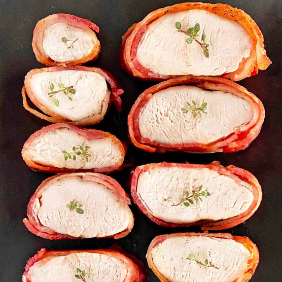 Sliced bacon wrapped turkey tenderloins on a black serving platter.