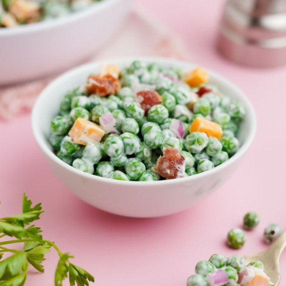 A small white bowl of creamy green pea salad with bacon and cheddar stands on a pale pink counter.