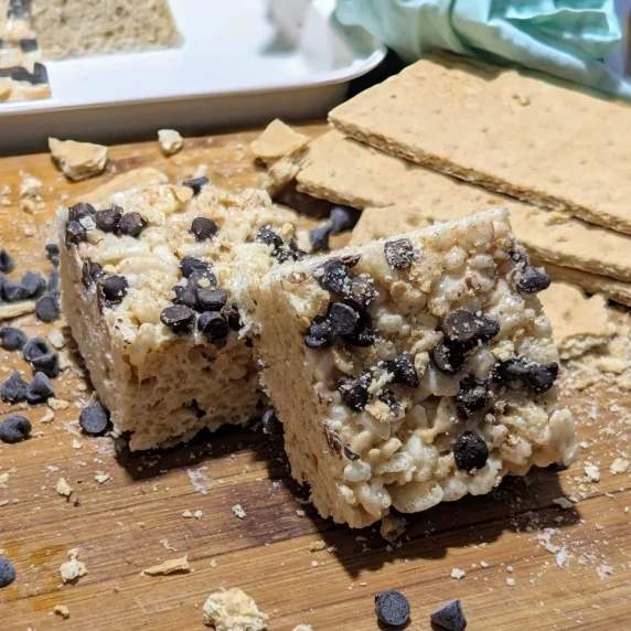 Two S'mores Rice Krispies Squares on a wooden board with a pile of graham crackers behind.