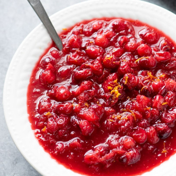 A spoon rests in a white bowl filled with homemade fresh cranberry sauce garnished with orange zest.