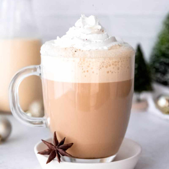 An eggnog latte topped with whipped cream and sprinkled with nutmeg in a glass mug on a white saucer