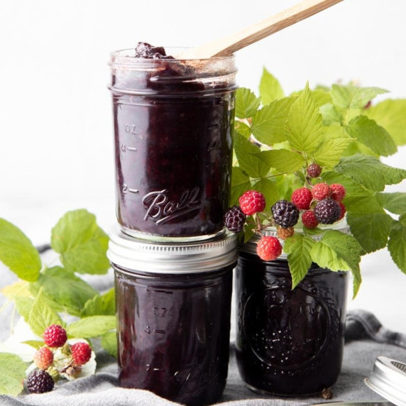 Three half-pint jars of black raspberry jam stacked with fresh raspberry sprigs around them.