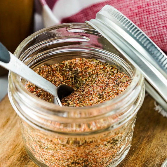 Side close up shot of steak rub in a glass jar with a tablespoon in it and the lid resting on side. 