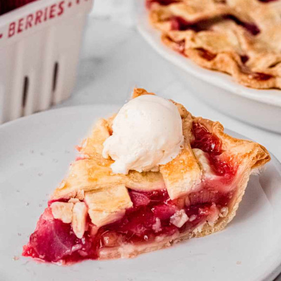 A lattice-topped slice of strawberry rhubarb pie with a scoop of vanilla ice cream on top on a plate