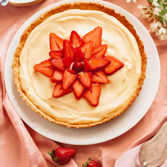 A French strawberry tart on a pink surface next to flowers and strawberries.